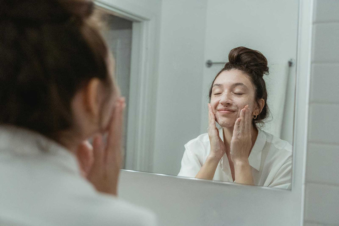 Young woman enjoying her skincare routine, gently washing her face with foam cleanser in front of the bathroom mirror.